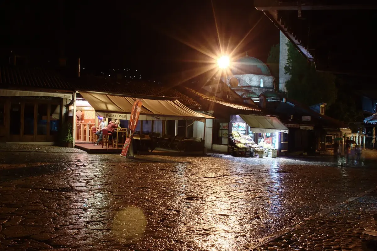 Baščaršija at night, with wet cobbles, a lit café and the dome of the Baščaršija mosque