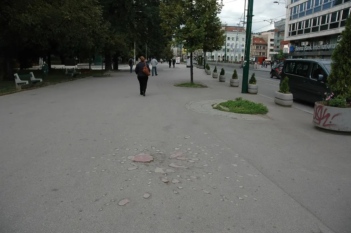 A Sarajevo Rose: a mortar scar filled with faded red resin in a city pavement, with people walking past